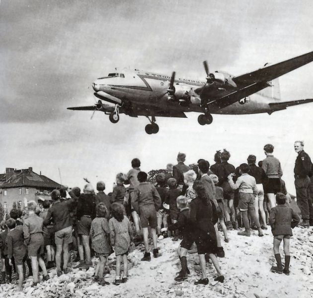 O fotografie istorică alb-negru realizată de jos în sus. În prim-plan, un grup mare de copii germani stă cocoțat pe un morman uriaș de dărâmături și moloz lăsat în urma războiului. Copiii sunt întorși cu spatele la cameră și privesc cu toții spre cer, fascinați. Deasupra lor, pe un cer parțial acoperit de nori, un avion militar masiv cu patru motoare, un Douglas C-54 Skymaster, este surprins în zbor la joasă înălțime, pregătindu-se să aterizeze. Pe fundal se zărește o parte din gardul aeroportului Tempelhof și siluetele unor clădiri urbane.