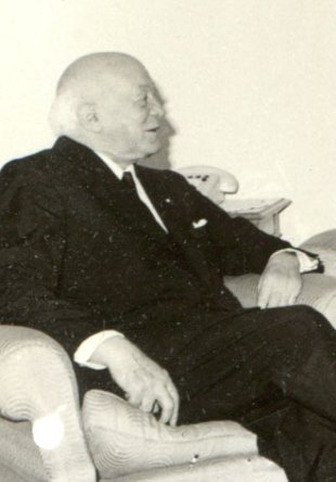 Fotografie istorică de epocă, realizată în plan mediu din profil, a savantului în vârstă Henri Coandă, îmbrăcat într-un costum negru elegant și așezat pe un fotoliu într-o încăpere.