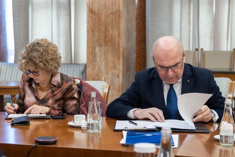 O fotografie de birou, captând un moment de colaborare intensă între prim-ministrul Ilie Bolojan și viceprim-ministrul Oana Gheorghiu. Ambii sunt așezați la o masă de lucru modernă, orientați unul spre celălalt, discutând și analizând împreună documente și un laptop deschis, subliniind o atmosferă de concentrare și dedicare în exercitarea atribuțiilor de stat.