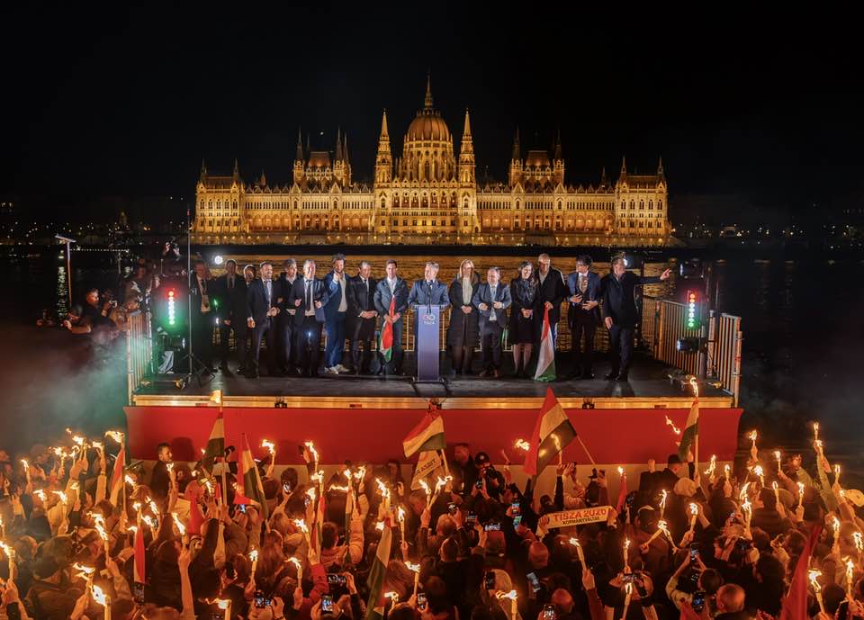 Vedere de noapte spectaculoasă a clădirii Parlamentului Ungariei, puternic luminată, în timp ce pe Dunăre, o mulțime imensă de susținători ai lui Peter Magyar, pe o platformă flotantă, țin torțe aprinse, creând un râu de foc și simbolizând dorința de schimbare.