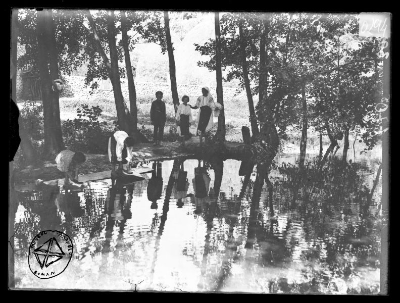 Fotografie alb-negru de epocă înfățișând mai multe persoane aflate pe malul unei ape, într-un cadru rural cu arbori și reflexii în apă.