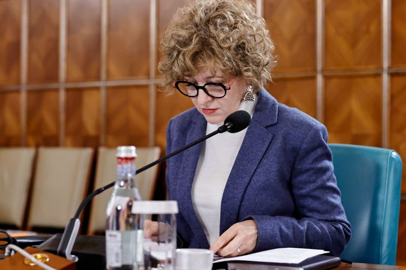 Oana Gheorghiu la o ședință oficială, citind documente la masa de lucru în fața unui microfon.