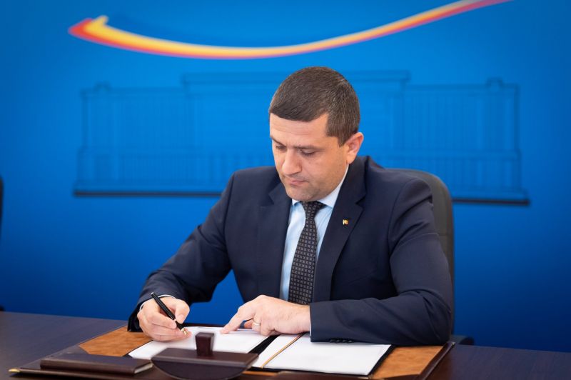 Ministrul Radu Miruță semnând un ordin oficial la birou. Acesta poartă un costum bleumarin și cravată, fiind așezat în fața unui fundal albastru cu logo-ul Guvernului României.