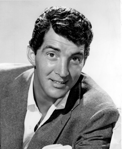 Portret fotografic alb-negru de epocă al lui Dean Martin, zâmbind larg, îmbrăcat într-un costum elegant și având o țigară aprinsă între degete.