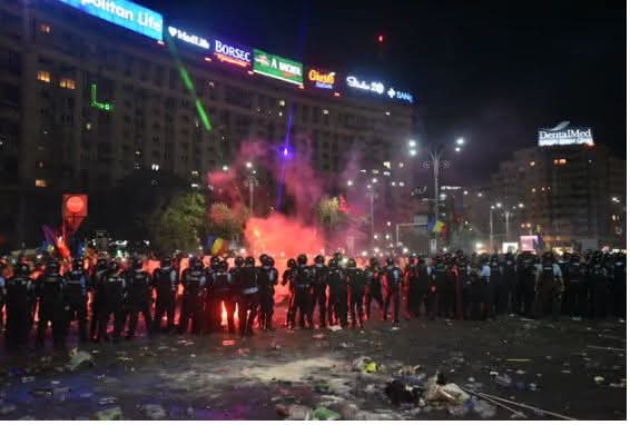 ​O mulțime compactă de protestatari pașnici în Piața Victoriei, București, în noaptea de 10 august 2018. În centrul imaginii se observă nori mari și denși de gaze lacrimogene lansate de jandarmi, care acoperă o parte a mulțimii în fața clădirii Guvernului.