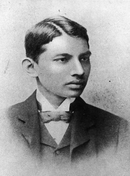 Portret fotografic alb-negru de epocă, tip bust, al tânărului Mahatma Gandhi purtând un costum occidental elegant cu vestă și papion.