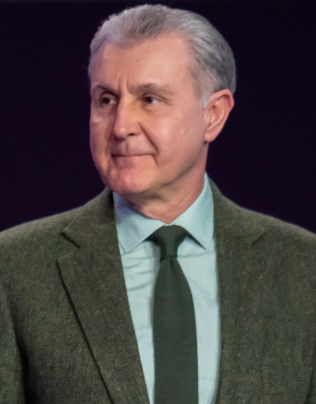 Portret fotografic color oficial de tip bust al Principelui Radu al României, îmbrăcat în costum închis la culoare, cămașă albă și cravată albastră, privind solemn spre cameră.