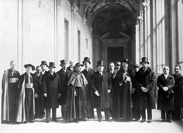 Fotografie istorică alb-negru în plan complet cu un grup mare de oficiali de la Vatican și politicieni italieni în haine de epocă, aliniați pe un coridor monumental cu tavan boltit.